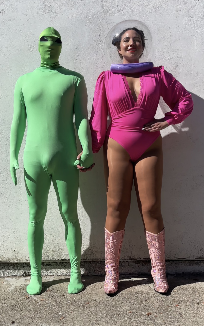 man in a green screen full body suit and an alien mask holding hands with a woman in a DIY Halloween costume of the 1985 Astronaut Barbie wearing a space helmet, pink bodysuit, and pink boots