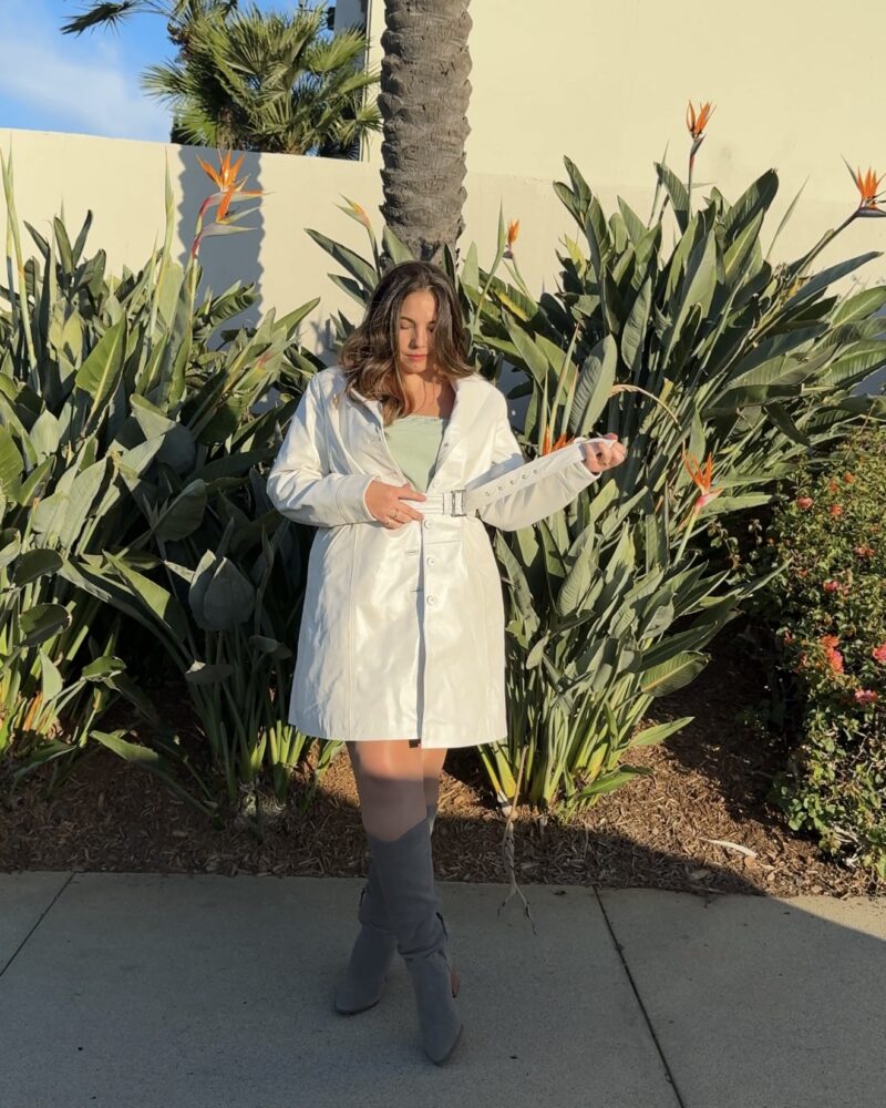 a woman (Lauryn Hock) wearing a fall outfit of tall suede boots and a trench coat leather jacket while standing in front of Birds of Paradise plants