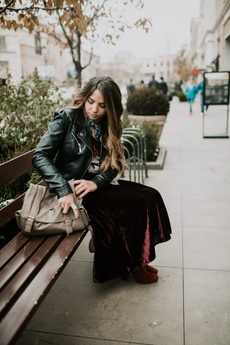 A young woman sits on a bench while looking through her purse.She's wearing a velvet skirt and a leather jacket