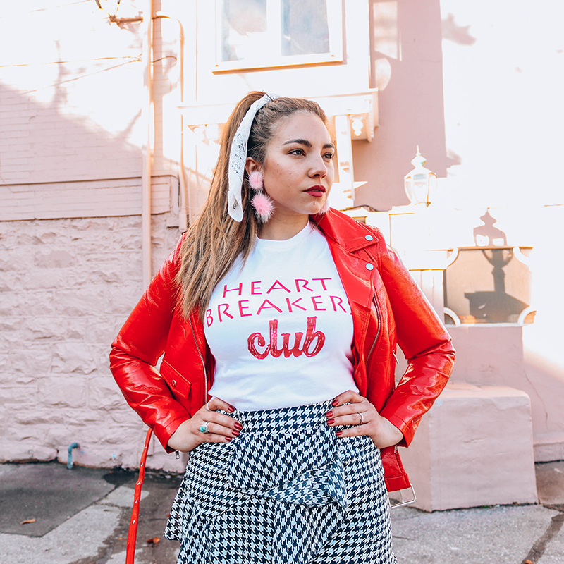 Lauryncakes wearing a Valentine's Day outfgit featuring a red leather jacket, gingham skirt, and a tee that says, "Heartbreakers Club" with her hands on her hips, a bow in her hair, and pom-pom earrings